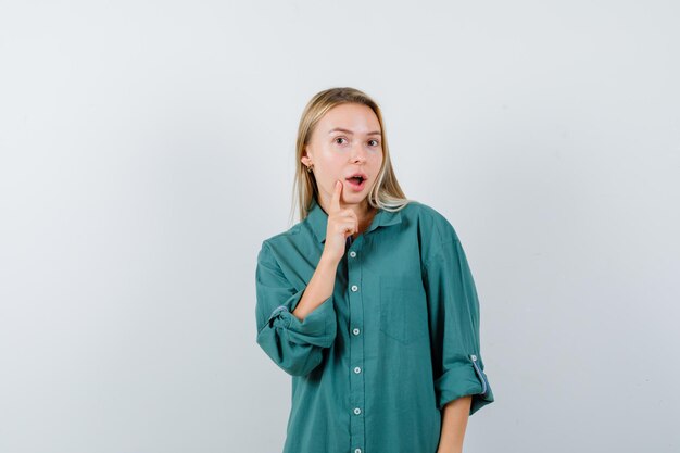 Blonde girl in green blouse putting index finger near mouth, opening mouth and looking surprised