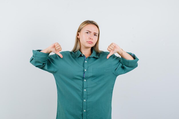Blonde girl in green blouse showing thumbs down with both hands and looking displeased