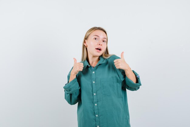 Blonde girl in green blouse showing thumbs up with both hands and looking pretty