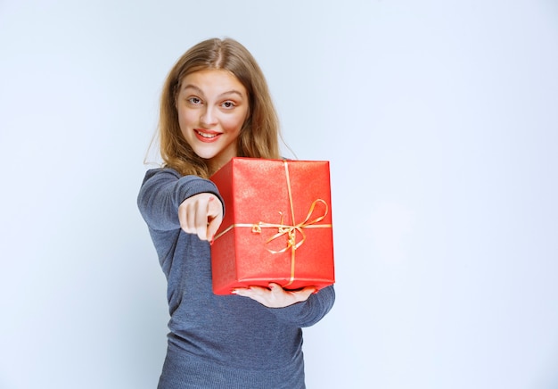 Бесплатное фото Блондинка девушка держит красную подарочную коробку и показывает кому-то.