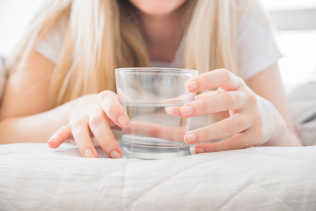 ブロンドの女の子がベッドの上の水のガラスを保持