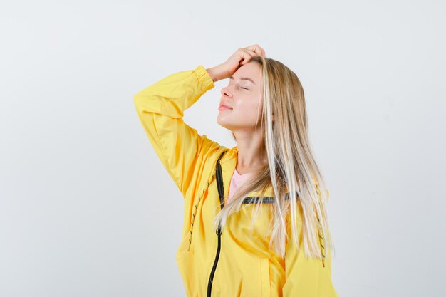 Blonde girl holding hand on head in yellow jacket and looking peaceful.