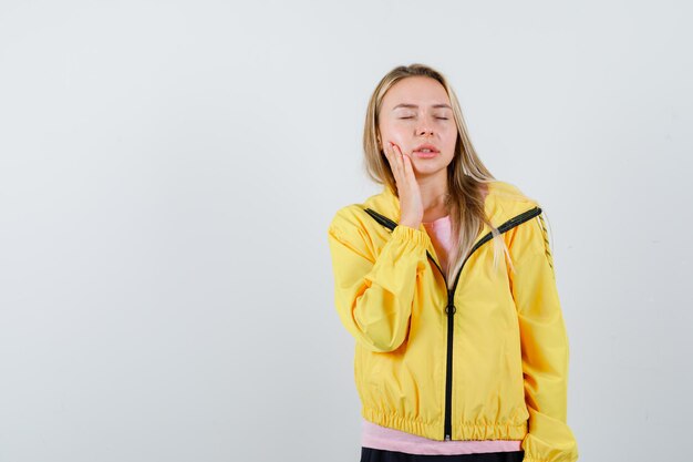 Blonde girl holding hand near mouth, having toothache in pink t-shirt and yellow jacket and looking exhausted