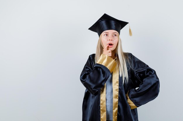 Blonde girl holding hand on waist while putting finger near mouth in graduation gown and cap and looking confident