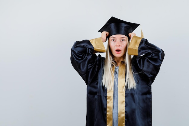 Blonde girl holding hands on cap, opening mouth in graduation gown and cap and looking surprised
