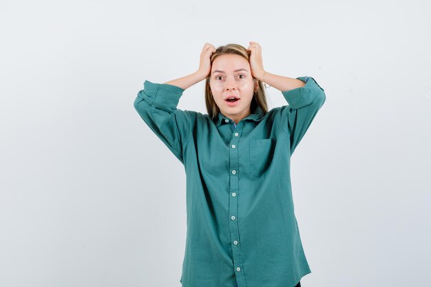 Blonde girl holding hands on head in green blouse and looking surprised.