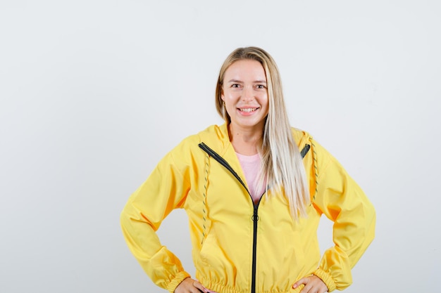 Бесплатное фото Блондинка девушка держит руки на талии в желтой куртке и выглядит веселой.