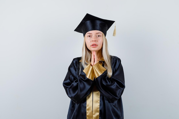 Blonde girl holding hands in prayer position in graduation gown and cap and looking serious