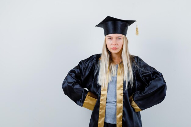 Blonde girl holding hands on waist in graduation gown and cap and looking confident