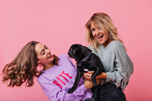 blonde girl holding her dog and laughing. Indoor portrait of refined ladies having fun together and playing with puppy.