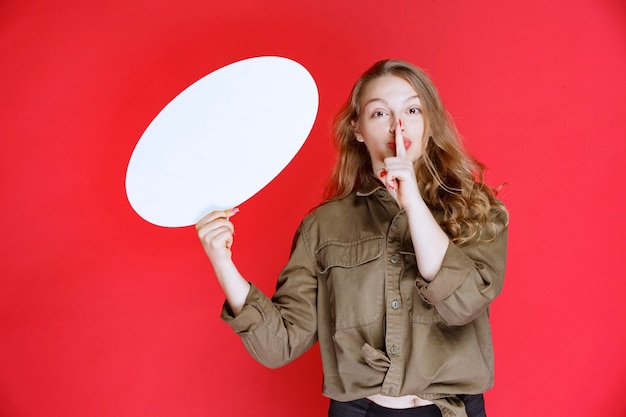 Foto gratuita ragazza bionda che tiene una lavagna ovale e chiede silenzio.