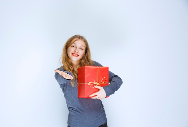 Blonde girl holding a red gift box and showing someone.