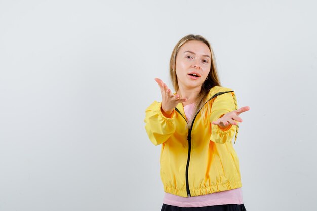 Blonde girl inviting to come in pink t-shirt and yellow jacket and looking amiable