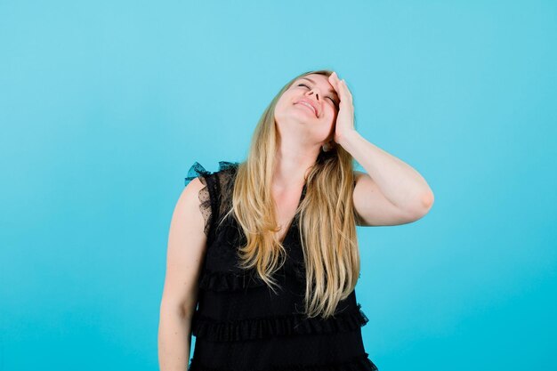 Blonde girl is closing eyes and putting hand on foreehad on blue background