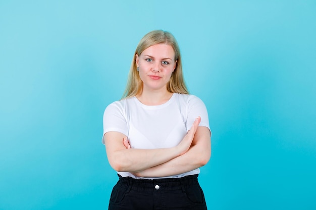 Blonde girl is looking at camera by crossing arms on blue background