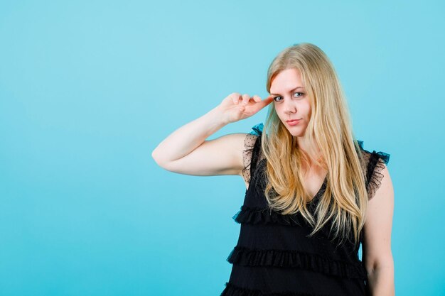 Blonde girl is looking at camera by holding forefinger on temple on blue background