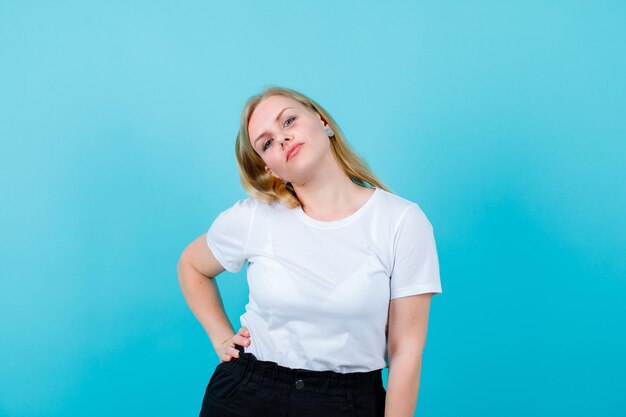 Blonde girl is looking at camera by putting hand on waist on blue background