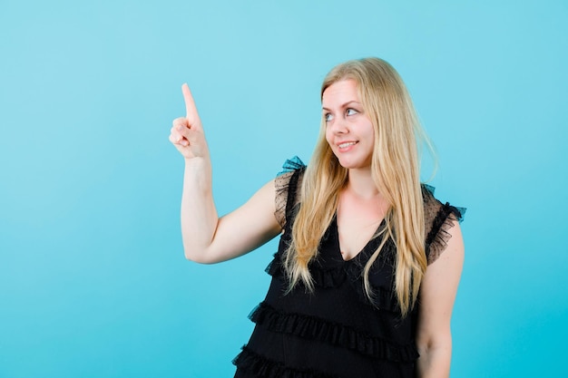 Blonde girl is looking and pointing up with forefinger on blue background