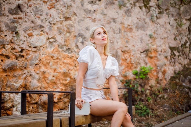 Blonde girl is looking up by sitting on bench on stone background