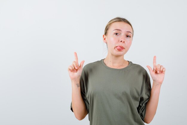 The blonde girl is pointing forefingers up by showing tongue on white background