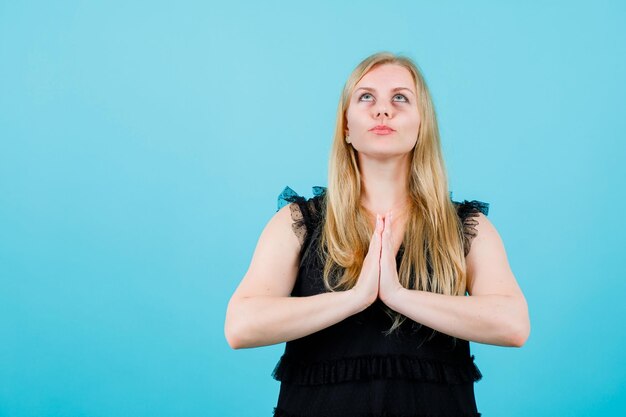 Blonde girl is praying by holding hands together and looking up on blue background