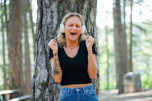 Blonde girl is screaming and raising up her fists while listening music on headphone on nature background