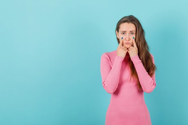 Blonde girl is showing sad face mimicry by holding forefingers on cheeks on blue background