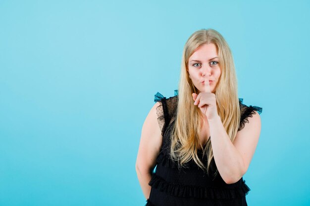 Блондинка показывает жест молчания, держа указательный палец на губах на синем фоне