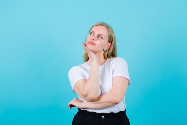 Blonde girl is thinking by putting hand under chin on blue background