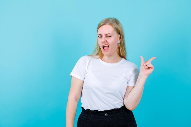 Blonde girl is winking and pointing rigth with forefinger on blue background