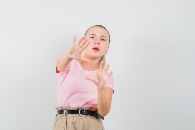 Blonde girl keeping hands to defend herself in t-shirt, pants and looking scared. front view.