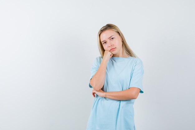 Free photo blonde girl leaning jowl on palm, standing in thinking pose in blue t-shirt and looking pensive , front view.