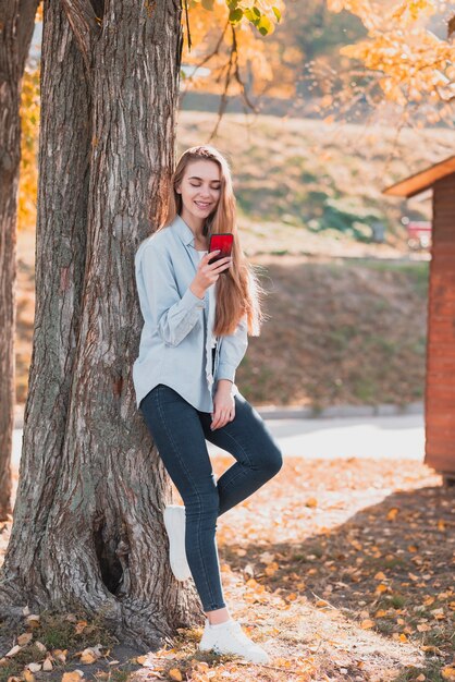 Блондинка смотрит на телефон и сидит рядом с деревом
