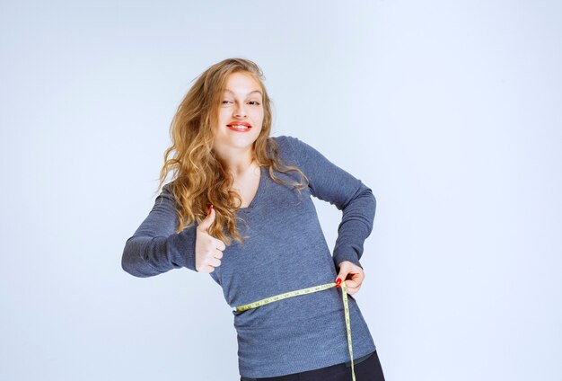 Blonde girl measuring her waist size with a tape and feeling successful.