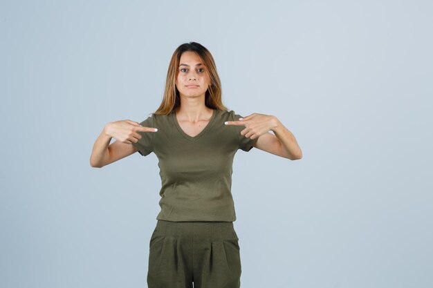 Foto gratuita ragazza bionda in maglietta verde oliva e pantaloni che si punta con le dita indice e sembra affascinante, vista frontale.