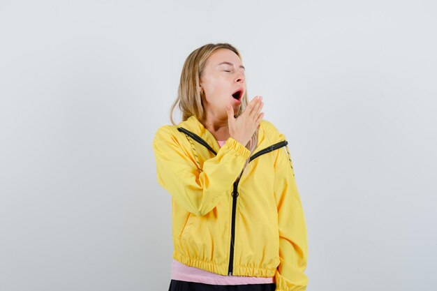 Блондинка в розовой футболке и желтой куртке держит руку возле рта, зевает и выглядит сонной