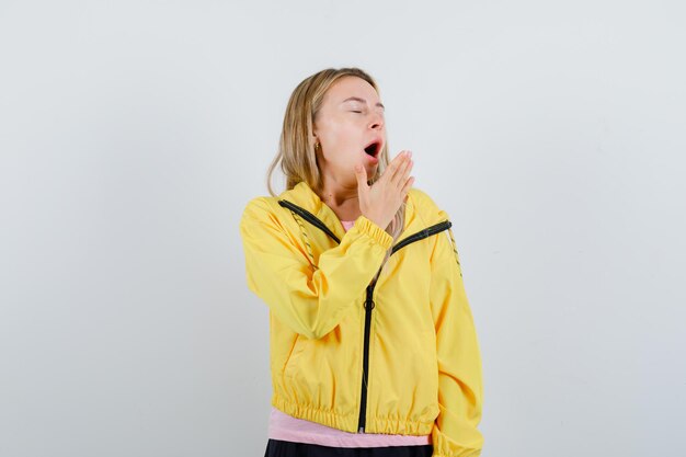 Blonde girl in pink t-shirt and yellow jacket holding hand near mouth, yawning and looking sleepy