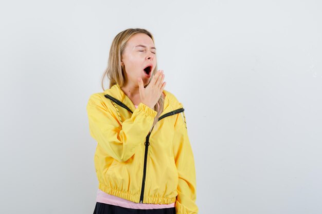 Blonde girl in pink t-shirt and yellow jacket holding hand near mouth, yawning and looking sleepy