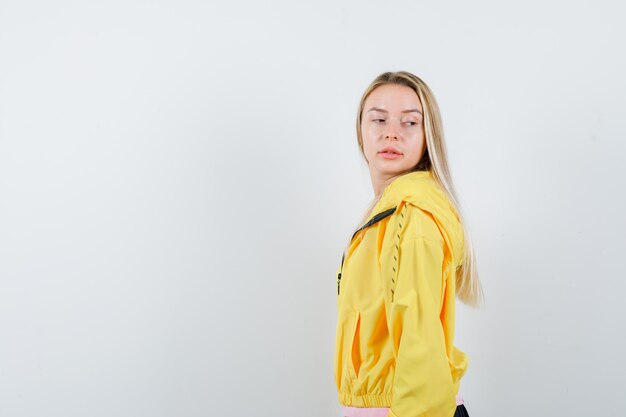 Blonde girl in pink t-shirt and yellow jacket looking over shoulder while posing at camera and looking alluring