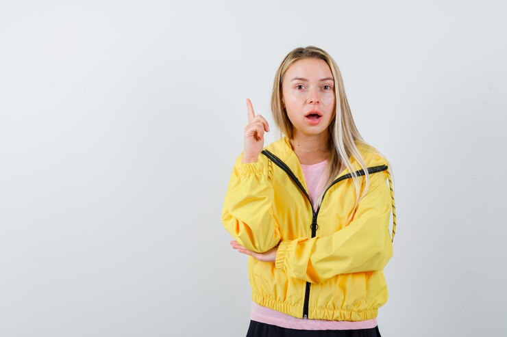 मुफ़्त फोटो blonde girl in pink t-shirt and yellow jacket raising index finger in eureka gesture while holding hand on elbow and looking sensible