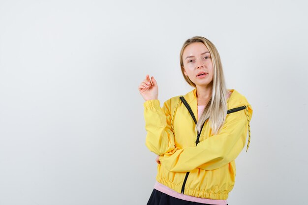 Blonde girl in pink t-shirt and yellow jacket raising one hand in eureka gesture while holding hand on elbow and looking alluring