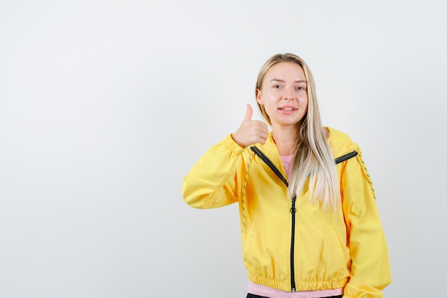 Blonde girl in pink t-shirt and yellow jacket showing thumb up and looking satisfied