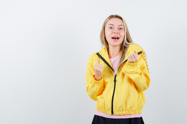 Blonde girl in pink t-shirt and yellow jacket showing winner gesture and looking happy