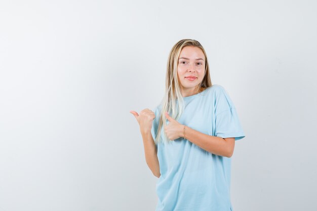 Blonde girl pointing aside with thumbs in blue t-shirt and looking cheery , front view.