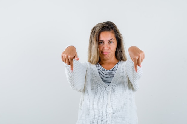 Blonde girl pointing down with index fingers in striped t-shirt, white cardigan and jean pants and looking happy. front view.