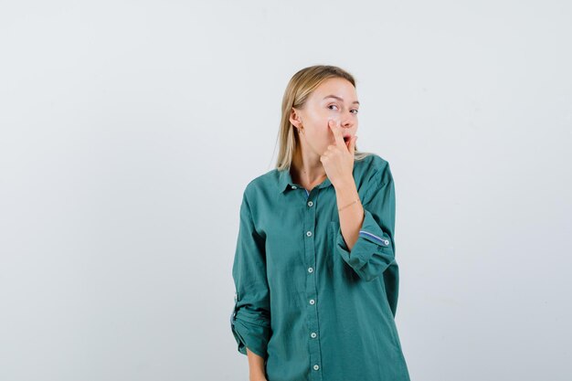 Blonde girl pointing face with index finger in green blouse and looking focused.