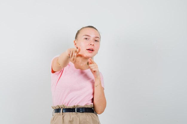 Tシャツ、パンツで正面を指して、陽気に見えるブロンドの女の子、正面図。