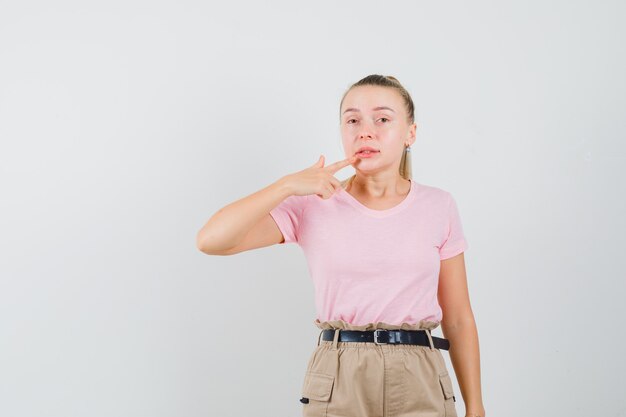 Blonde girl pointing at her lips in t-shirt, pants front view.