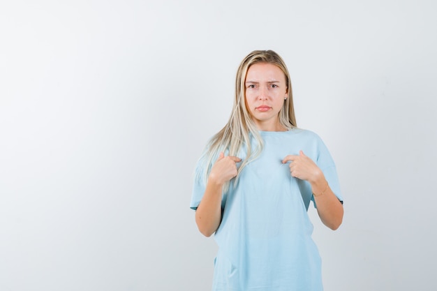 Blonde girl pointing at herself with index fingers in blue t-shirt and looking serious. front view.