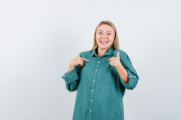 Blonde girl pointing at herself with index fingers in green blouse and looking happy